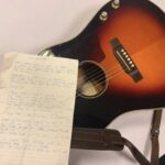 Photo showing an acoustic guitar with a handwritten lyric sheet over the fretboard
