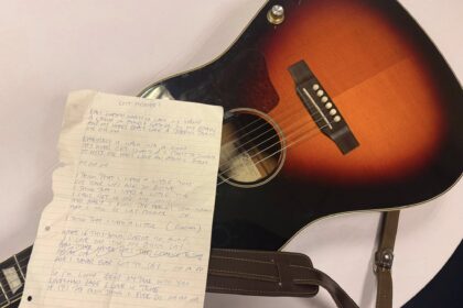 Photo showing an acoustic guitar with a handwritten lyric sheet over the fretboard